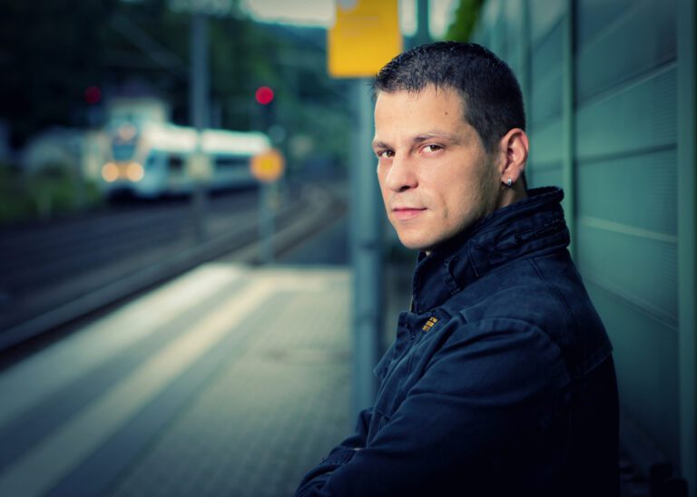 Das Foto zeigt einen jungen Mann mit Jacke auf einem Bahnhof, nur der Oberkörper und der Kopf sind zu sehen.