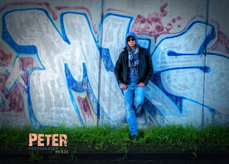 Das Foto zeigt einen jungen Mann in Lederjacke und mit Cap auf, vor einer Betonwand mit Graffiti.