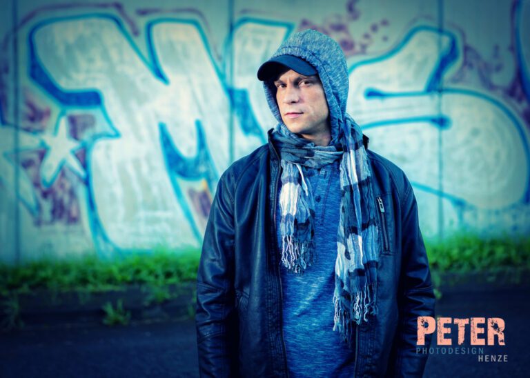 Das Foto zeigt einen jungen Mann in Lederjacke und mit Cap auf, vor einer Betonwand mit Graffiti.