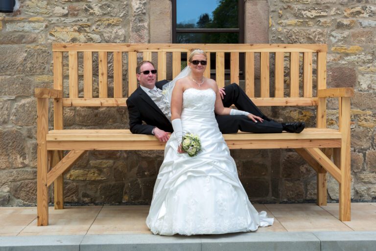 Das Foto zeigt ein Ehepaar an bzw. auf einer sehr großen Holzbank, beide mit Sonnenbrillen