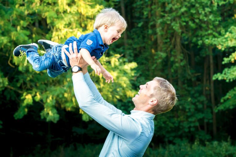 Das Foto zeigt einen jungen Mann, der seinen ganz jungen Sohn nach oben wirft und ihn wieder auffängt. Beide lachen sich an.