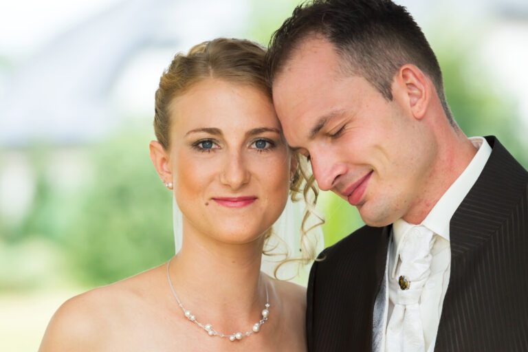 Das Foto zeigt ein junges Ehepaar. er hat seinen Kopf an ihre Schläfe gelehnt und hat die Augen geschlossen, sie schaut in die Kamera.