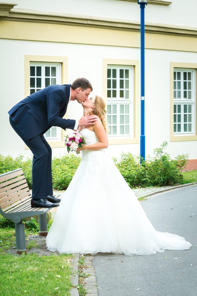 Das Foto zeigt ein junges Ehepaar. Er steht auf einer Parkbank und küsst seine Frau von oben herunter.